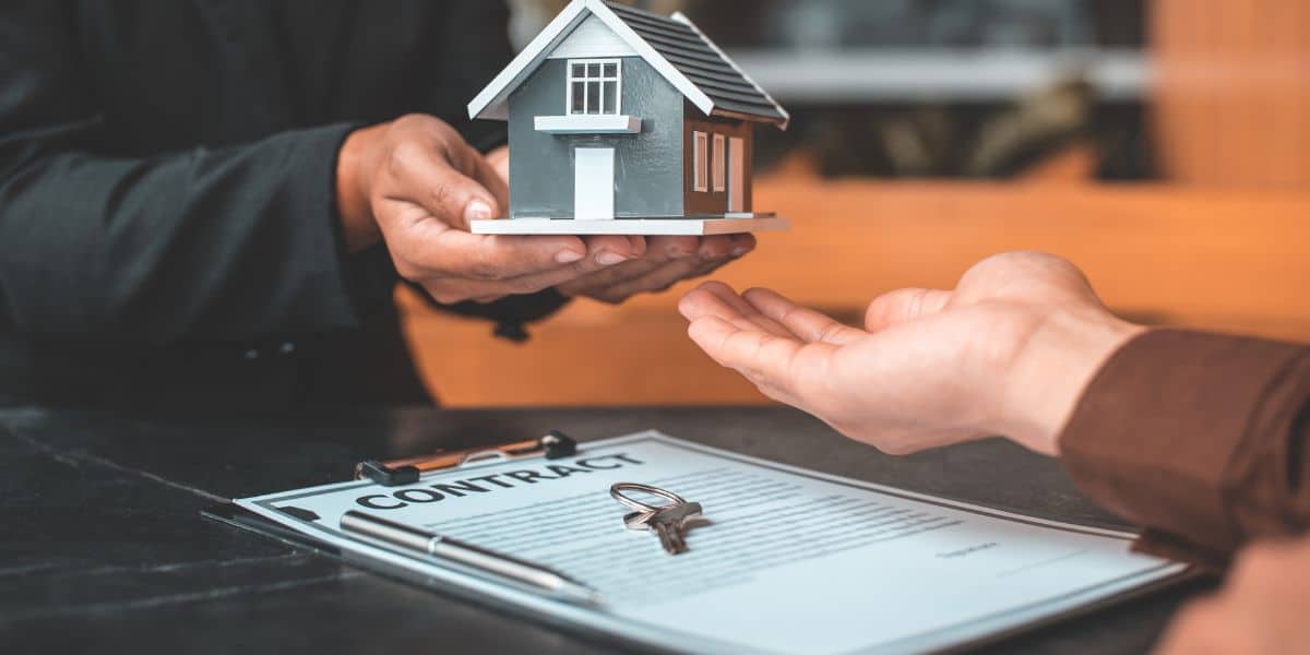 Person handing over a small house model above a contract with keys on the table.