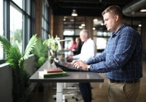 Confident real estate franchise owner stands in a bright, modern office, looking thoughtfully out a large window at the ci...